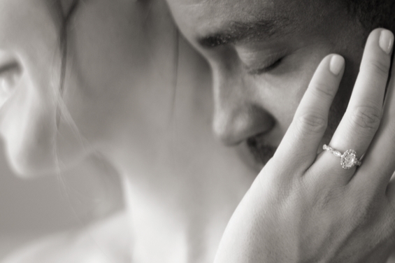 Black and white image of a bride&amp;s hand on the groom&amp;s cheek, showing off her engagement ring with oval diamond center.